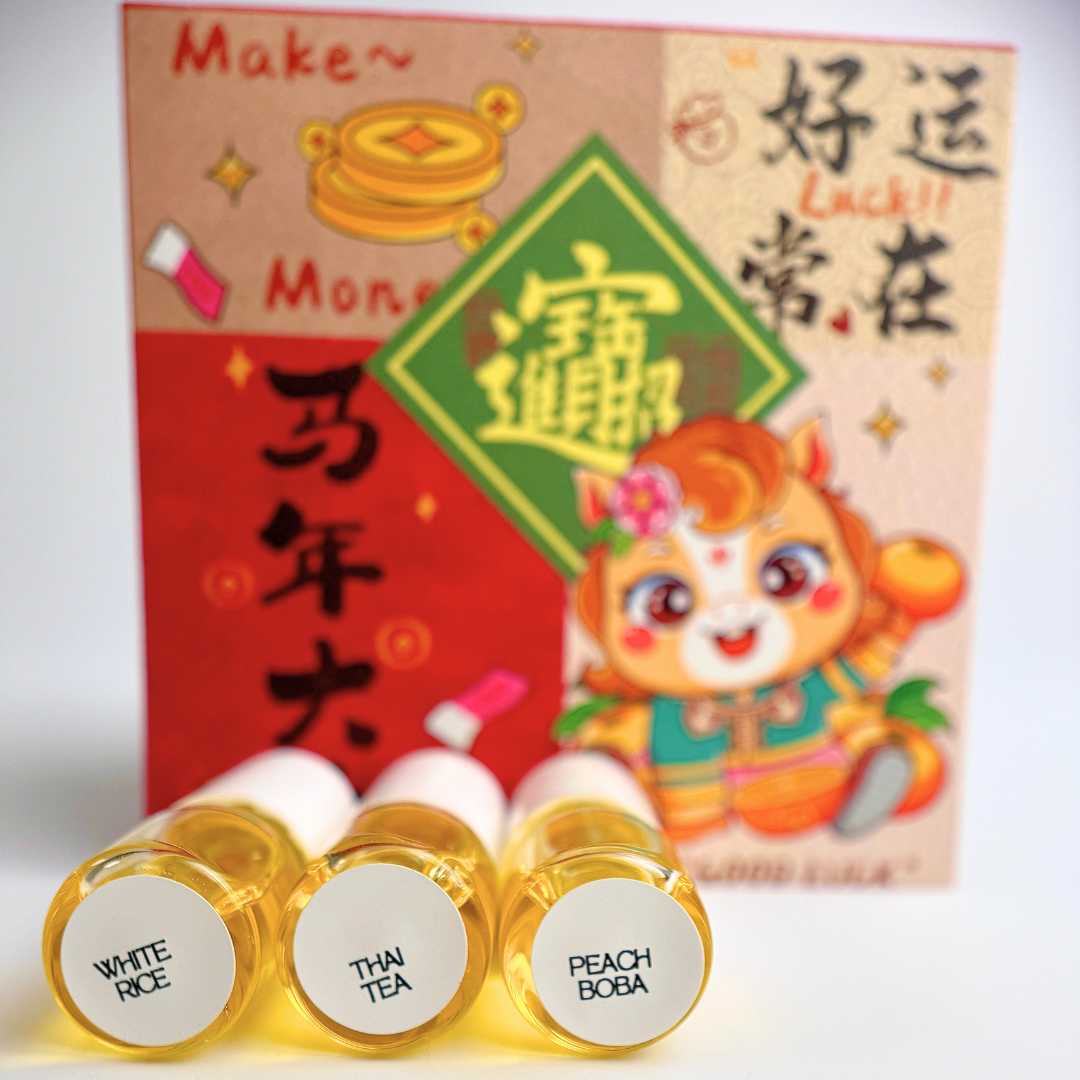 Three small bottles of cuticle oil labeled "white rice", "thai tea" and "peach boba" in front of a colorful red envelope featuring a cartoon horse and Chinese text.