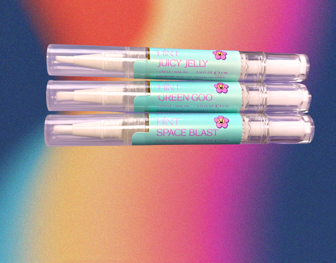 Three cuticle oil tubes laying down against a colorful gradient background. The tubes are clear plastic with light blue labels featuring pink text and small pink flower graphics.