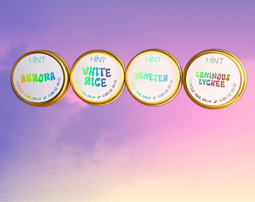 Four round tins of HINT brand cuticle balm arranged in a row against a pastel purple and pink gradient background. The tins are labeled Aurora, White Rice, Demeter, and Luminous Lychee.
